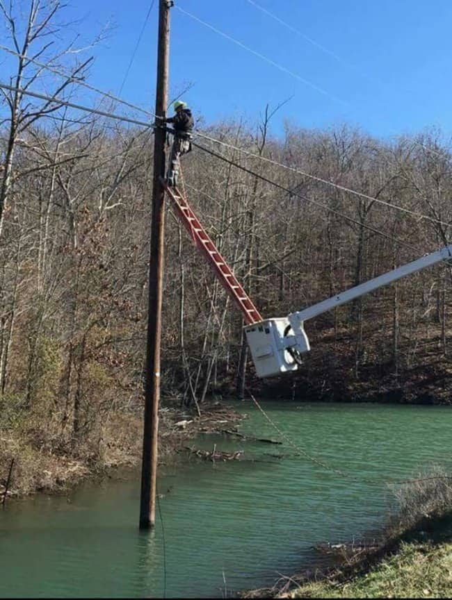 Lineman climbing a utility pole standing in water as a bucket truck reaches from the bank
