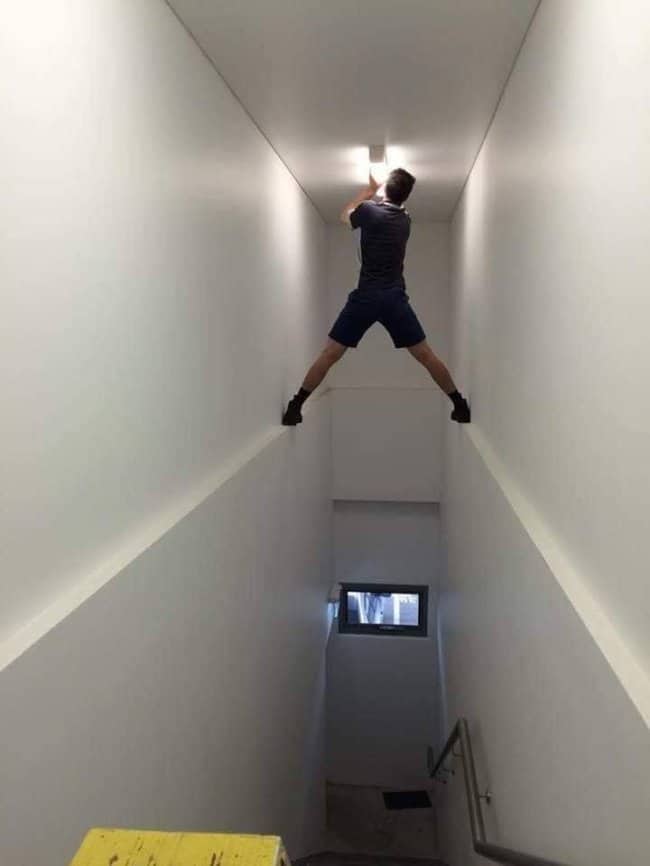 Man wedged between two stairwell walls with his legs spread wide to reach a ceiling light