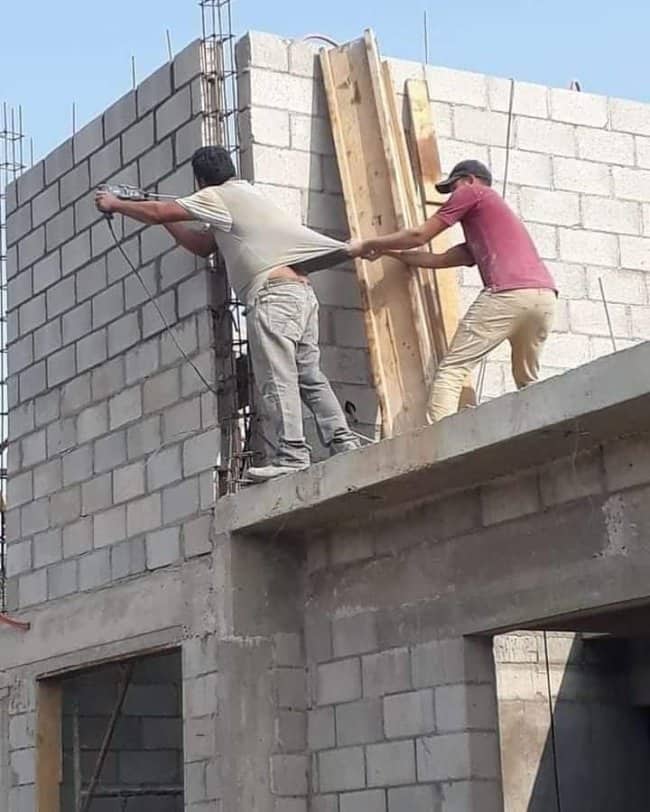 Two workers on an unfinished concrete roof edge, one leaning backward held only by a belt
