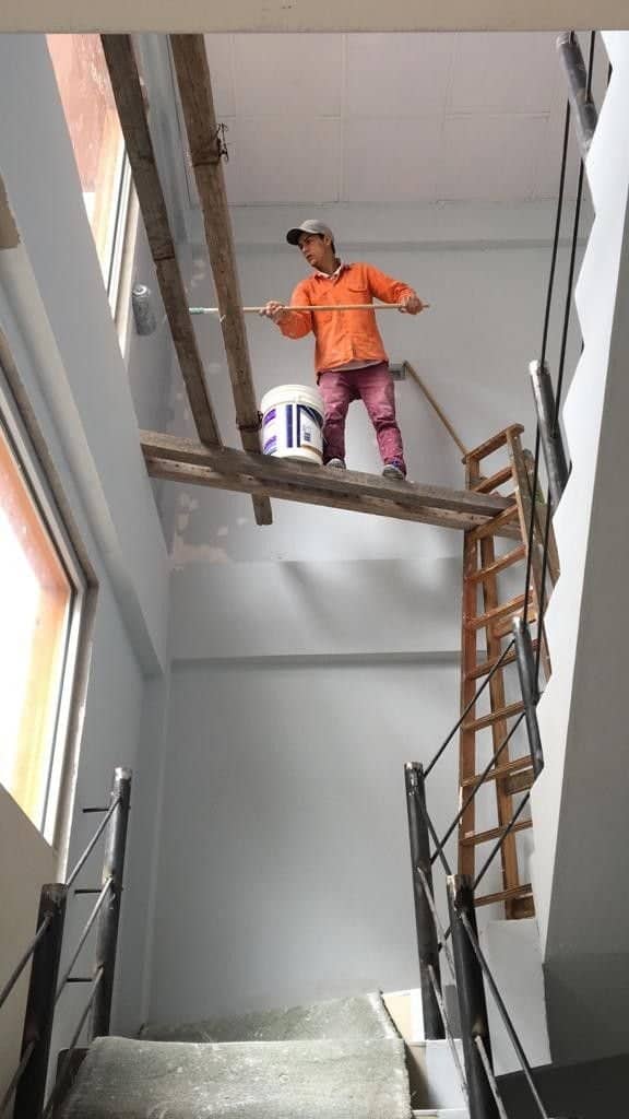 Painter standing on a narrow wooden plank suspended across an open stairwell holding a roller