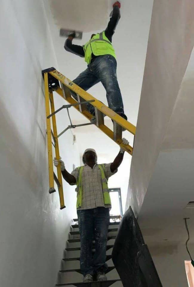 Worker on a stepladder balanced on a staircase while a colleague holds one leg up
