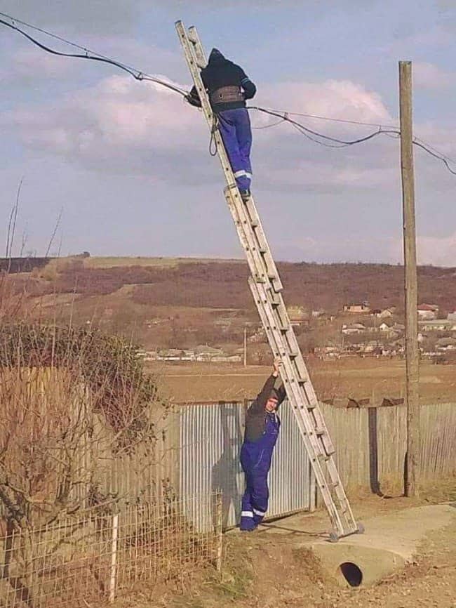 Worker high on a ladder leaning against power lines while another man steadies the base