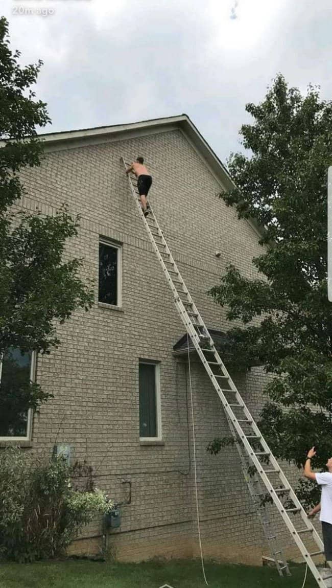 Shirtless man near the top of a tall extension ladder reaching a two story roofline