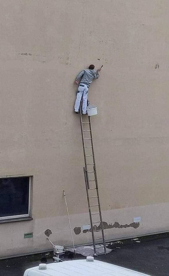 Painter perched at the top of a wooden ladder against a blank wall with a paint bucket