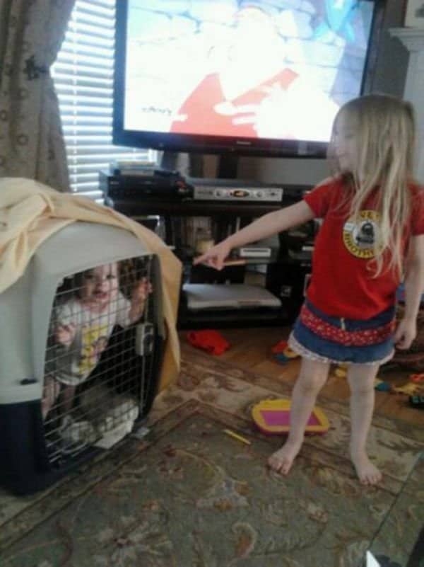 Young girl pointing at a toddler locked inside a pet crate indoors.