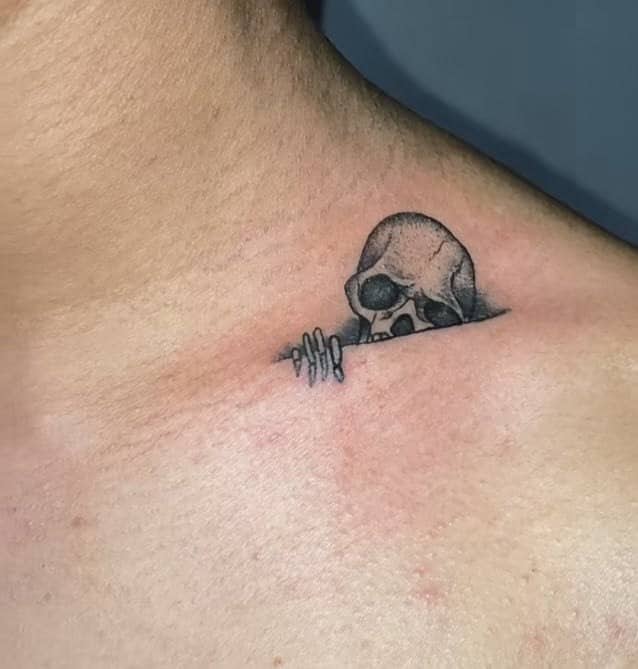 Small skeleton peeking over collarbone scar creating a humorously clever unique tattoo placement design.