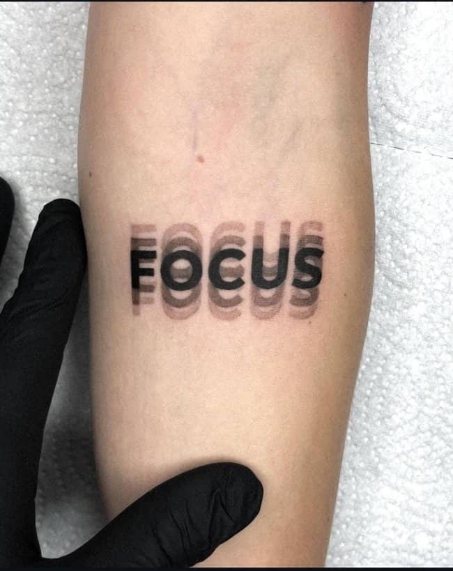 Blurry optical illusion text unique tattoo displaying the word focus with double vision effect.